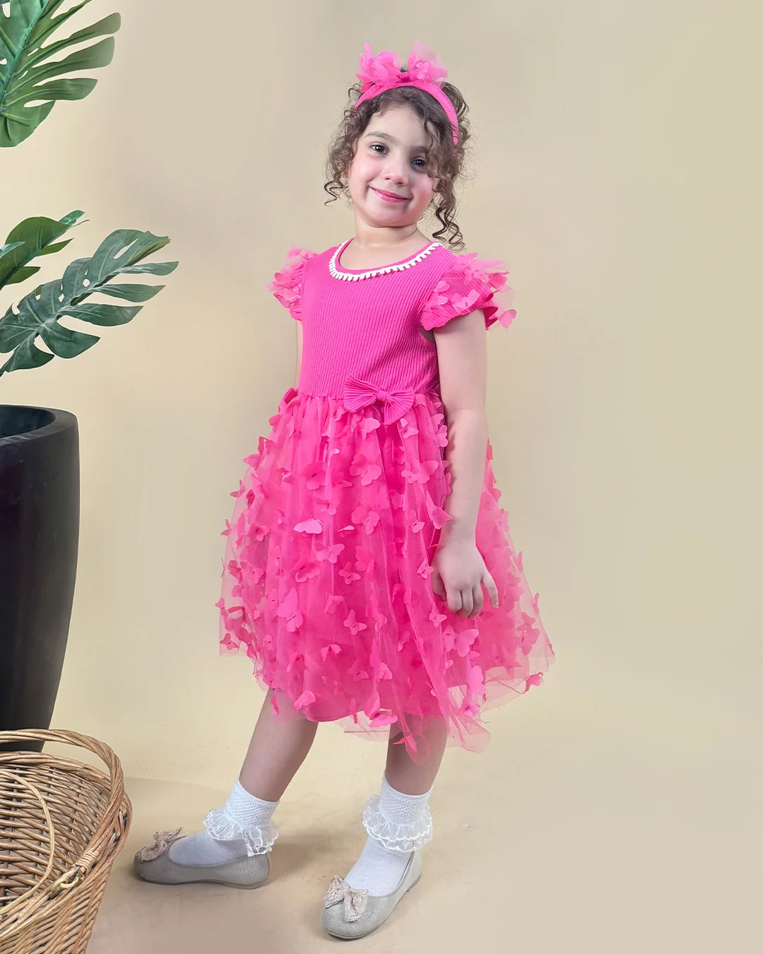 Young girl in a pink dress standing next to a plant on a beige background