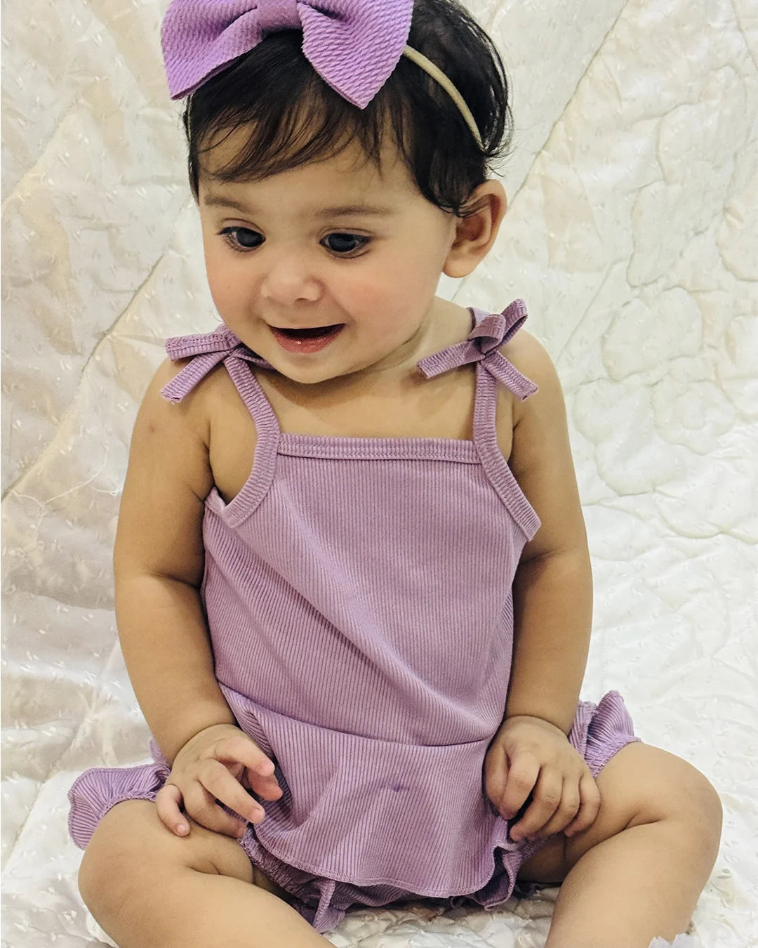 Baby wearing a purple outfit with a bow headband on a textured white background