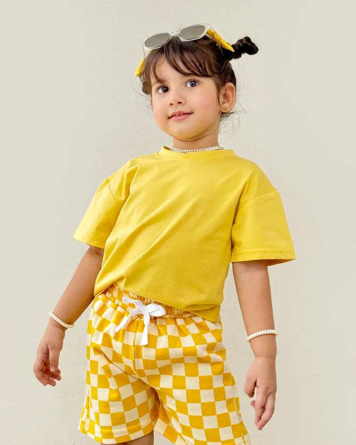 Child wearing a yellow outfit with checkered shorts on a plain background