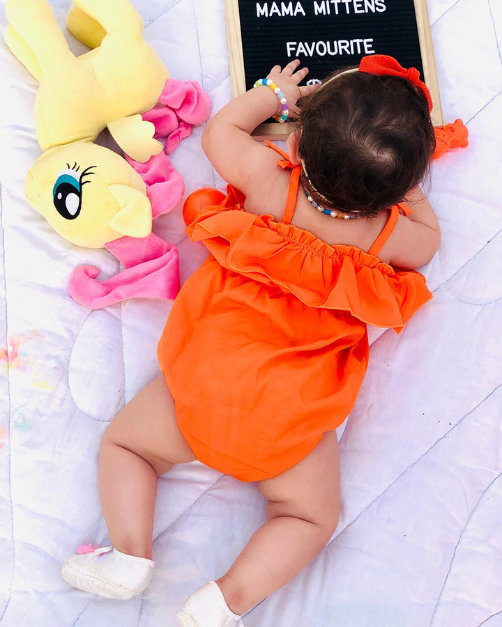 Baby in an orange outfit lying on a white blanket with colorful toys and a sign in the background.