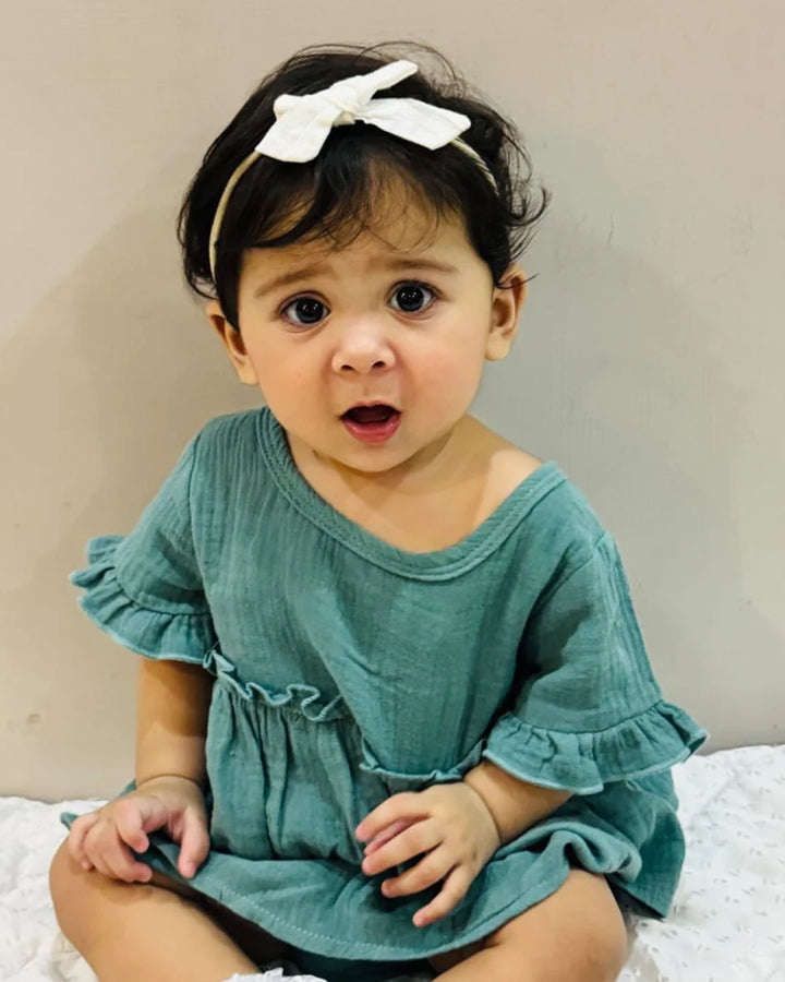 Child wearing a teal dress with ruffled sleeves and a white bow headband against a neutral background