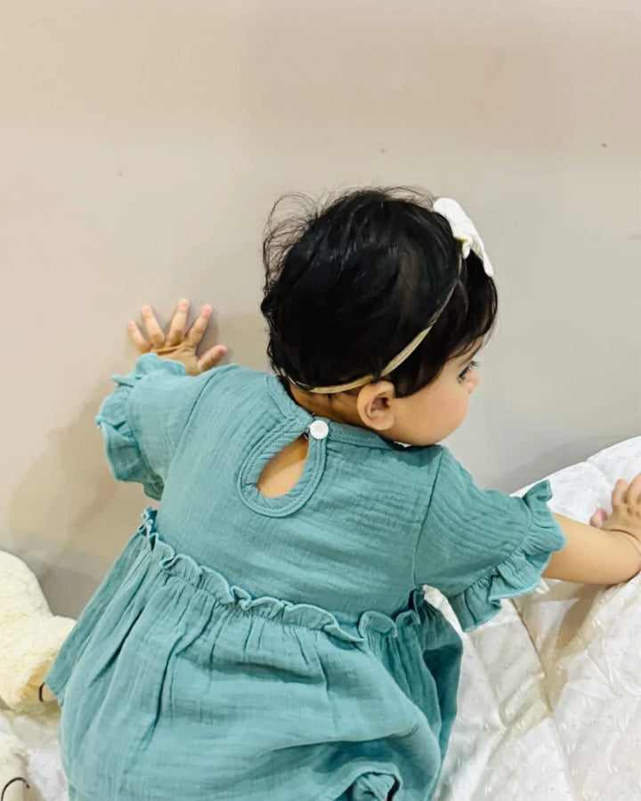 Child wearing a teal dress with ruffled sleeves and a white headband, sitting on a bed.