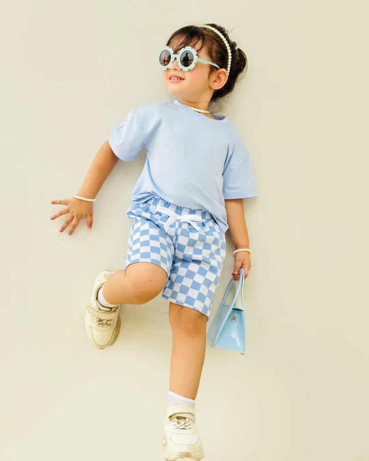 Child wearing a light blue outfit with checkered shorts, sunglasses, and holding a blue bag against a beige background.