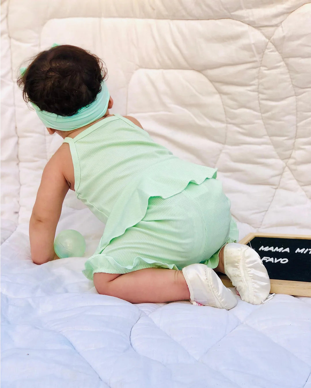 Baby in a green outfit sitting on a bed with a white quilt.