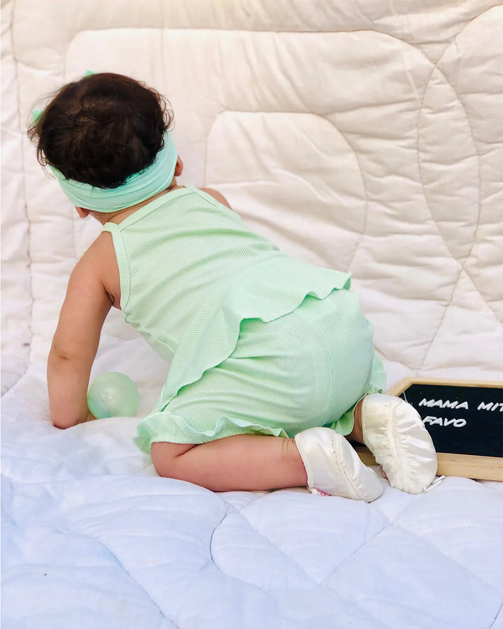 Baby in a green outfit sitting on a bed with a white quilt.