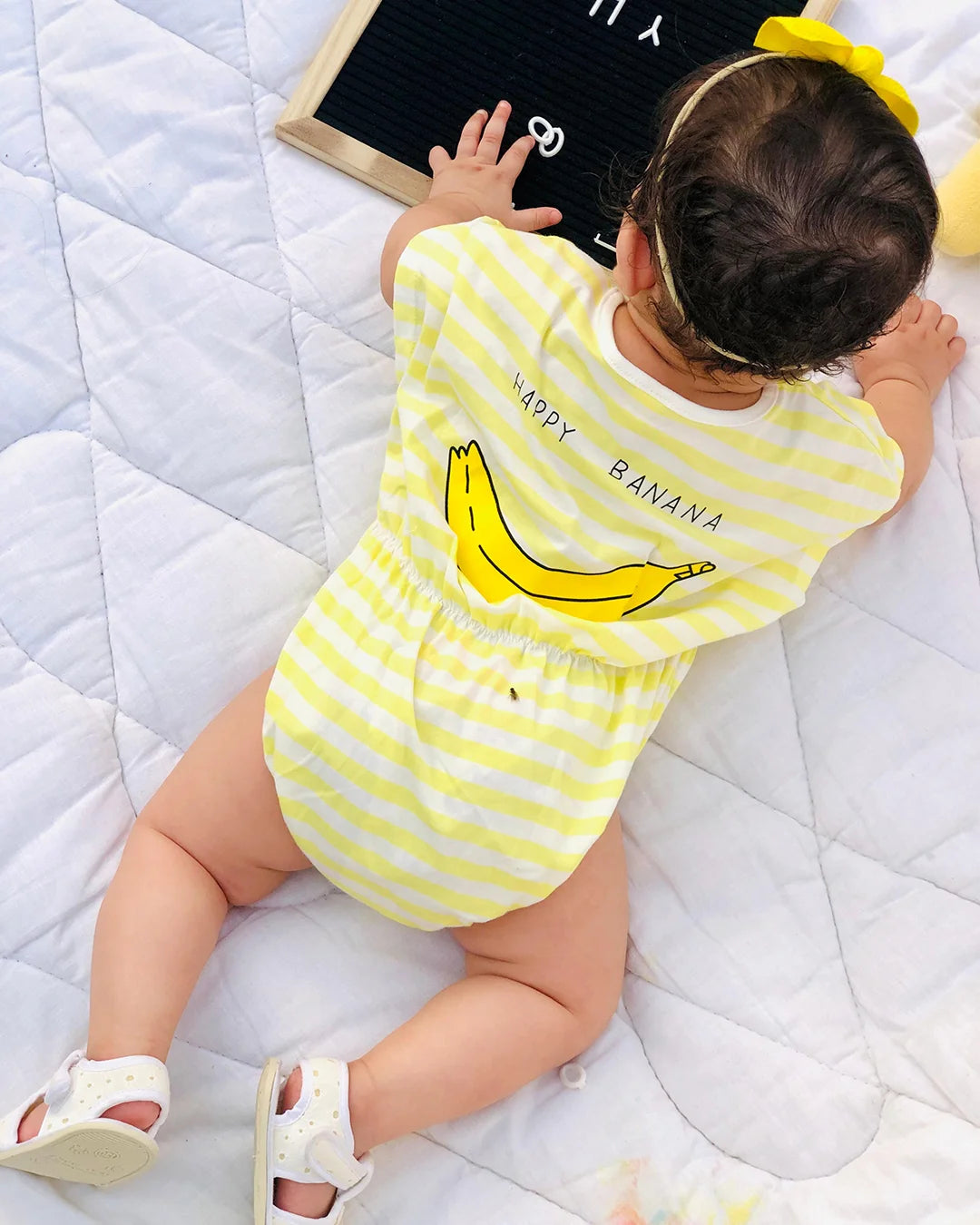 Baby wearing a yellow and white striped onesie with a banana design on a white blanket.