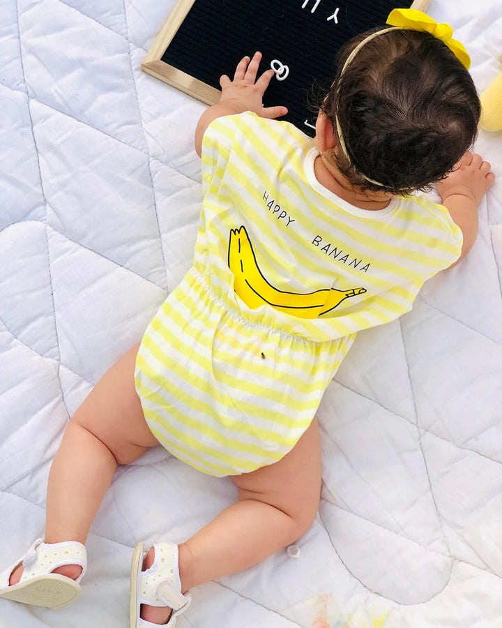 Baby wearing a yellow and white striped onesie with a banana design on a white blanket.