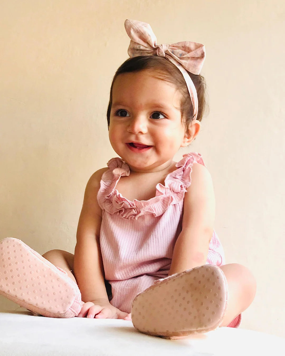 Baby in a pink outfit with a bow headband sitting on a light-colored surface.