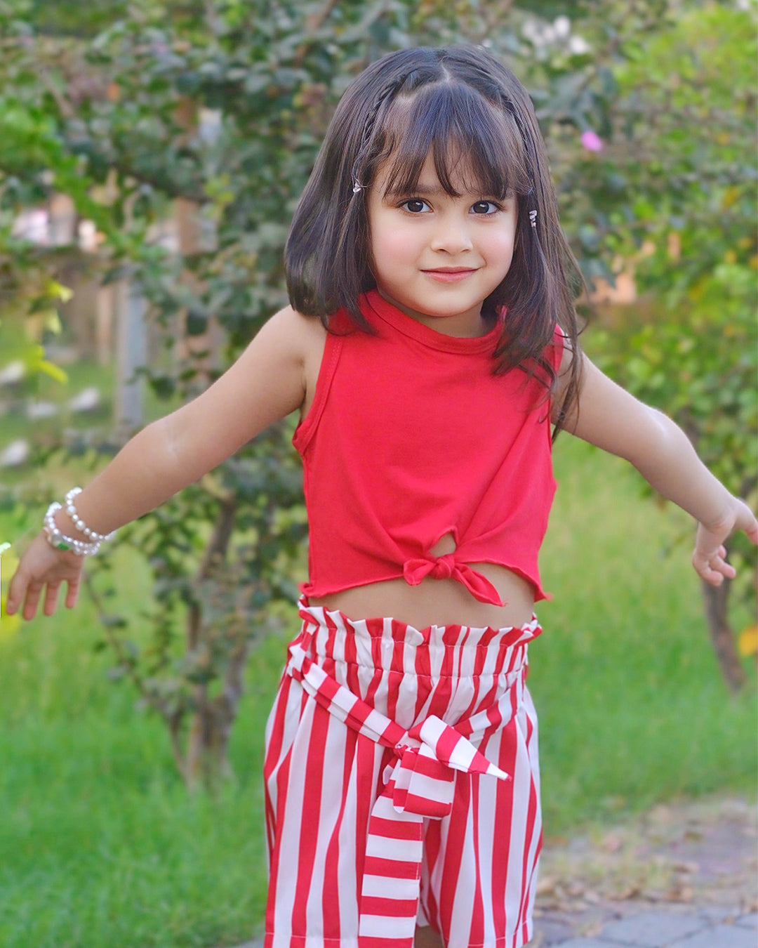 Young girl in a red top and red and white striped pants standing outdoors with greenery in the background