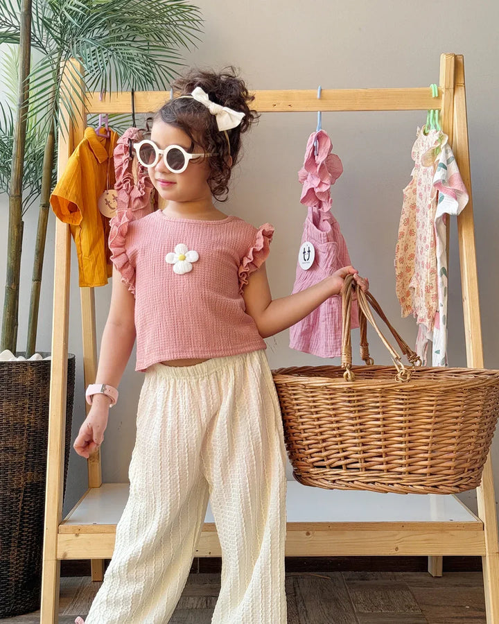 Child in a pink top and white pants holding a basket, standing next to a wooden rack with clothes.