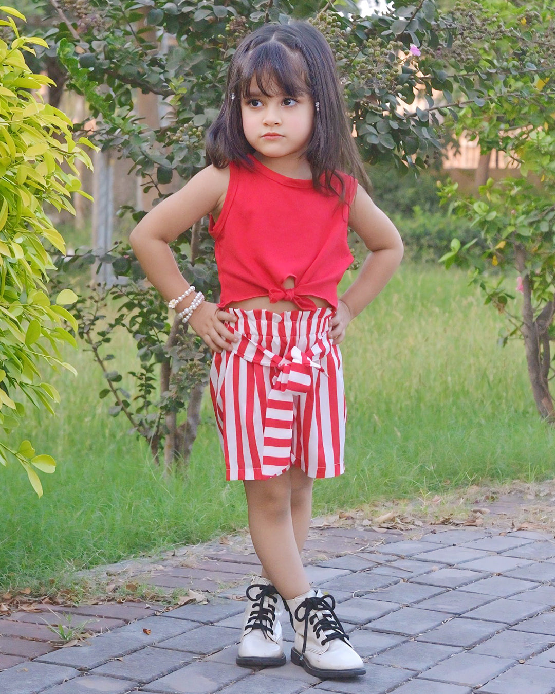 Child wearing a red top and red and white striped shorts standing on a paved path with greenery in the background.