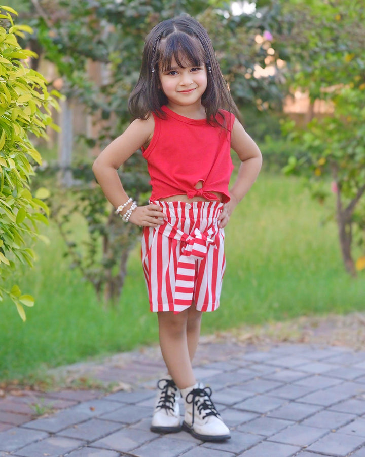 Child wearing a red top and striped shorts standing outdoors on a paved path with greenery.