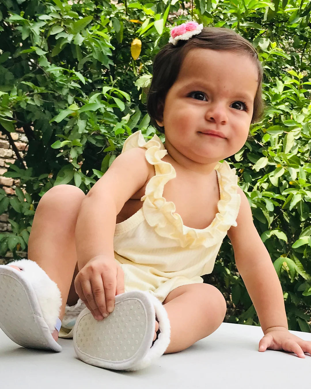 Baby in a yellow outfit with white shoes sitting outdoors among greenery