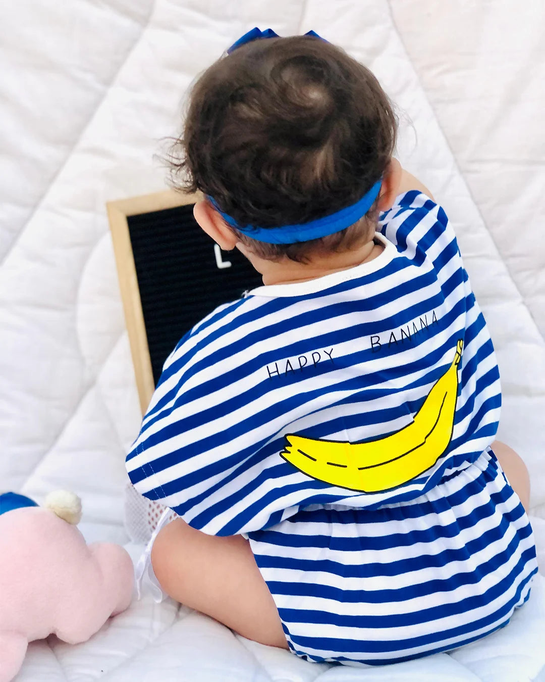 Child wearing a blue and white striped shirt with a banana graphic, lying on a white surface.