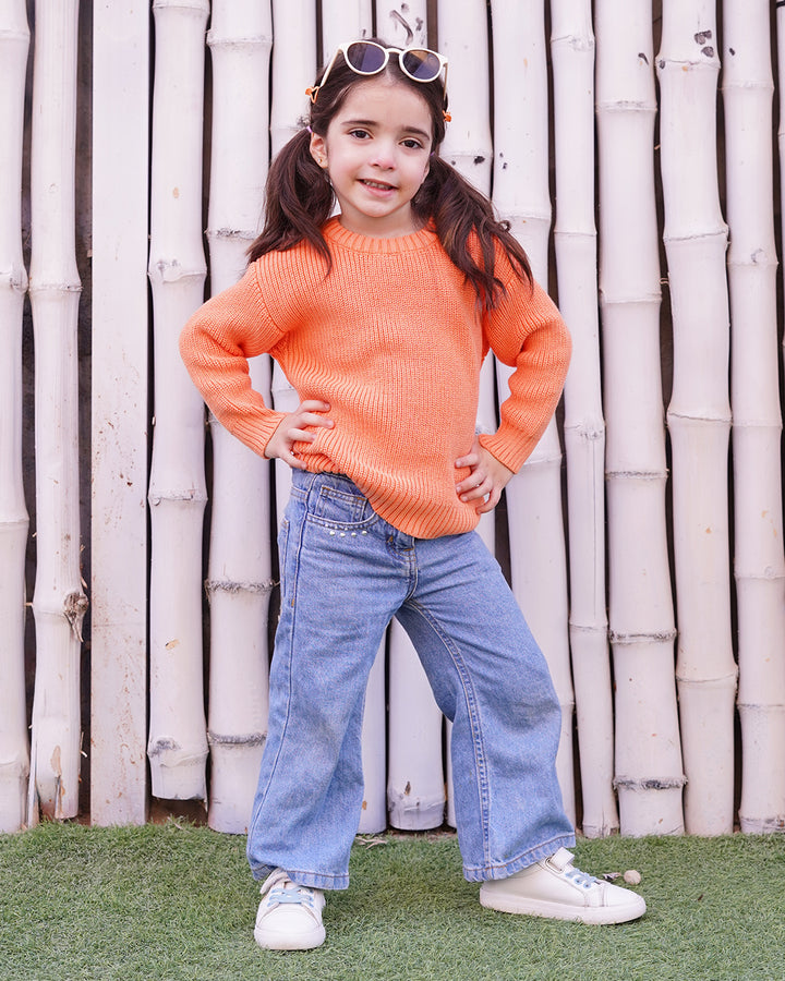 Young girl wearing an orange sweater and blue jeans standing in front of a bamboo fence.