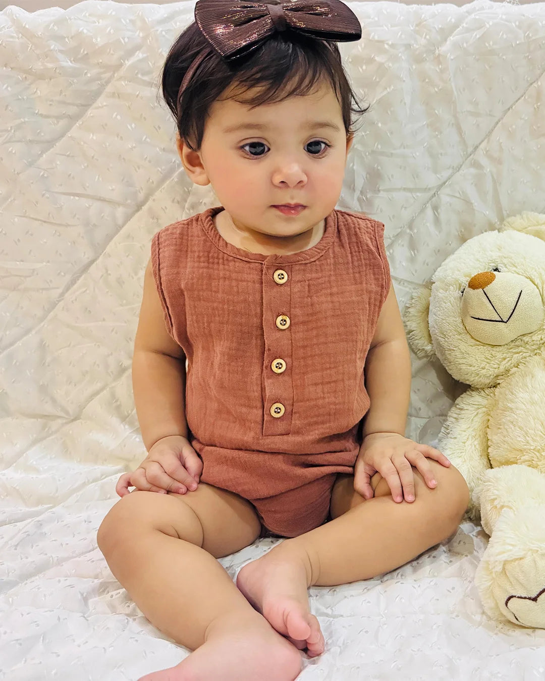 Baby in a rust-colored outfit with a bow sitting on a white blanket next to a teddy bear.