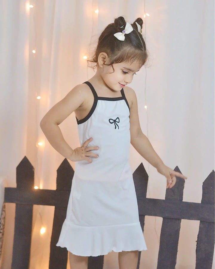 Young girl in a white dress with black trim standing in front of a decorative fence and light strings.