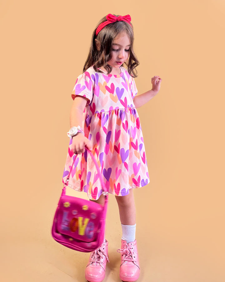 Young girl wearing a colorful heart-patterned dress holding a pink bag against a beige background
