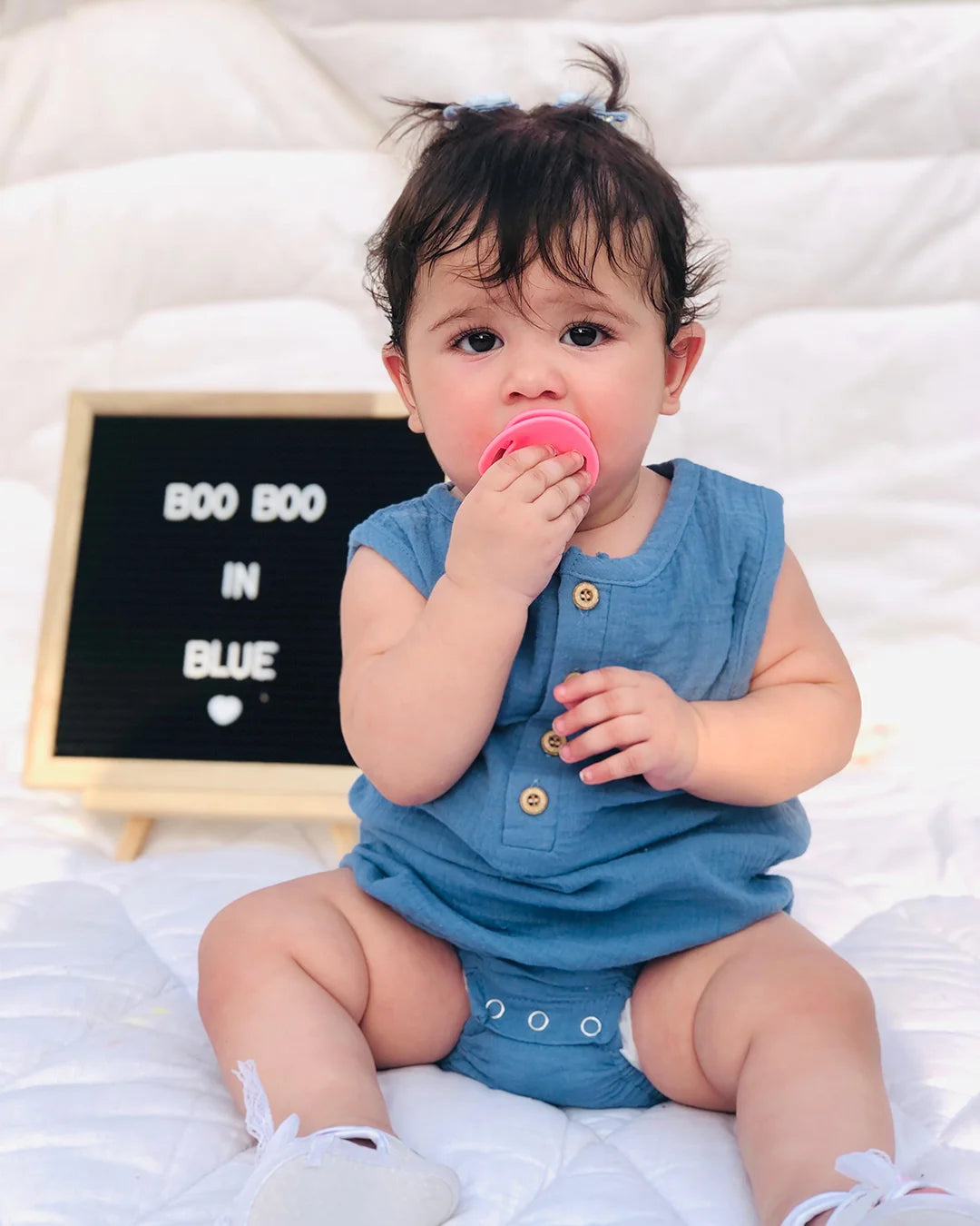 Baby in a blue romper holding a pink object, sitting on a white surface with a letterboard in the background.