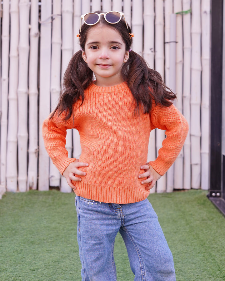 Young girl wearing an orange sweater and blue jeans standing outdoors.