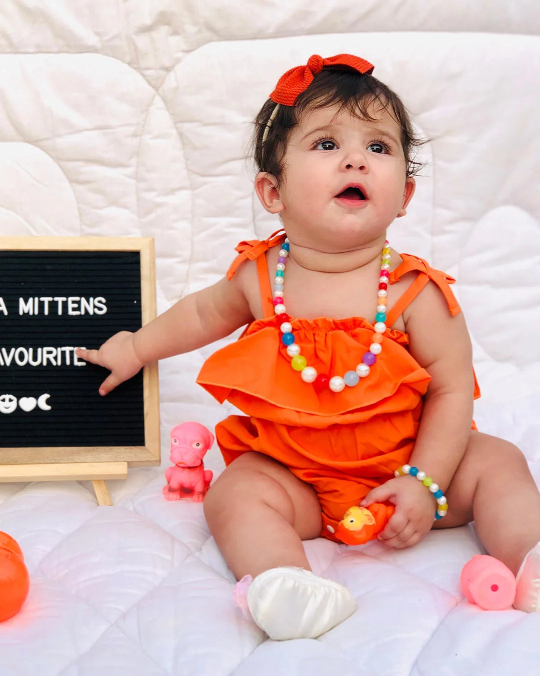 Baby in an orange outfit with colorful beads, sitting on a white blanket next to a blackboard.