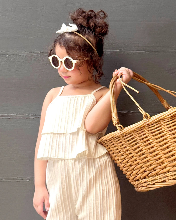 A young girl wearing a beige pleated cami top with spaghetti straps and a wide-leg pant co-ord set, accessorized with a headband and a woven basket.