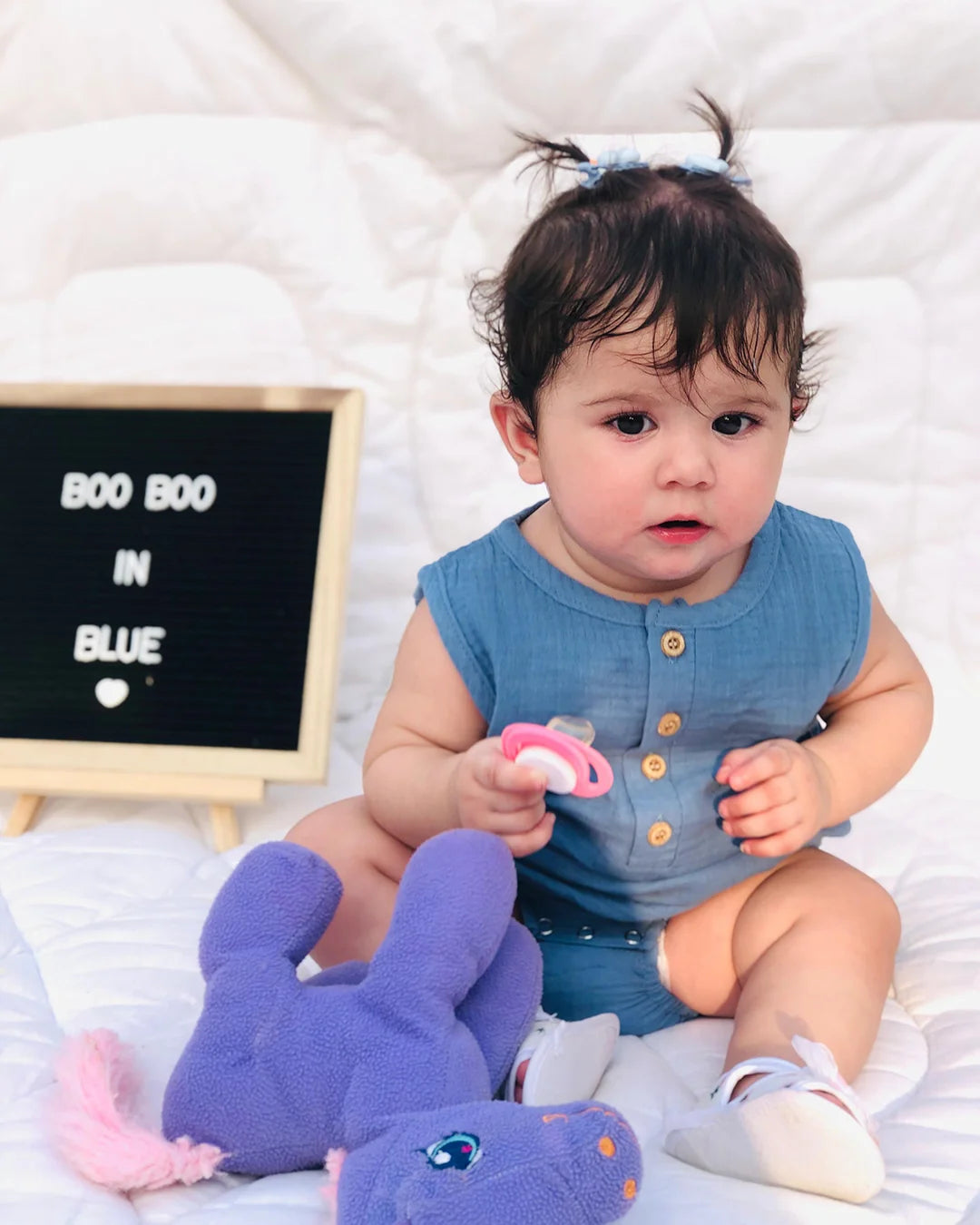 Baby in a blue outfit holding a pink pacifier, with a purple elephant toy and a sign in the background.