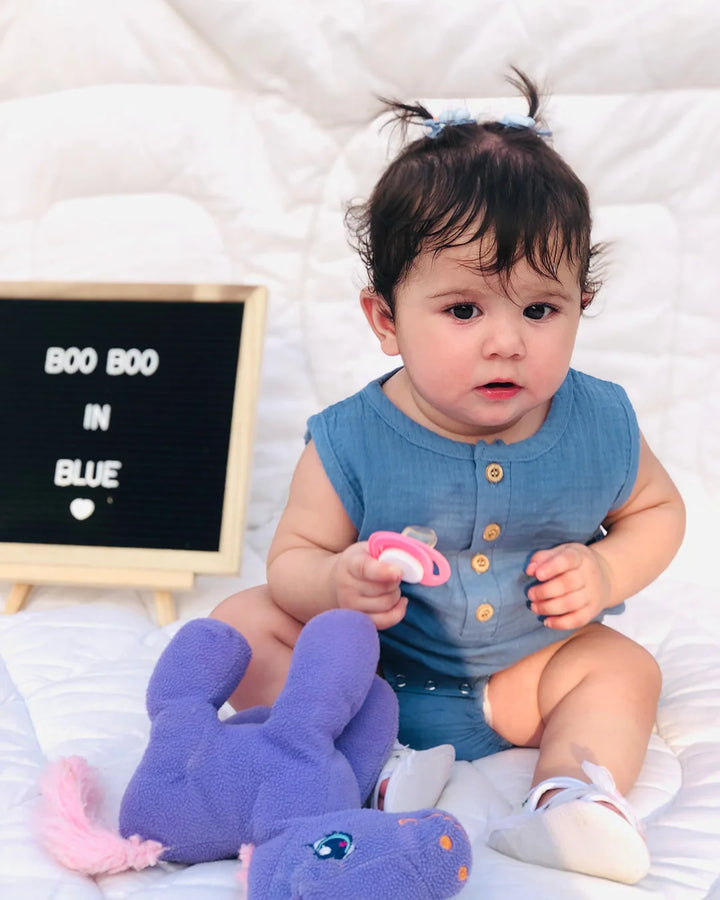 Baby in a blue outfit holding a pink pacifier, with a purple elephant toy and a sign in the background.