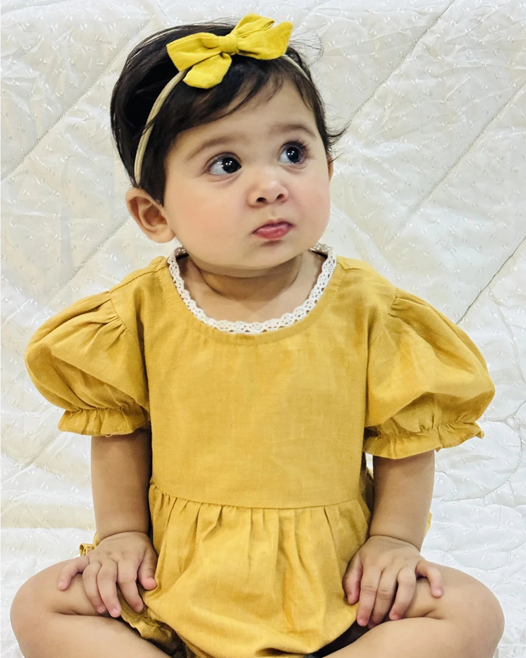 Child wearing a yellow dress and headband sitting on a white quilted surface
