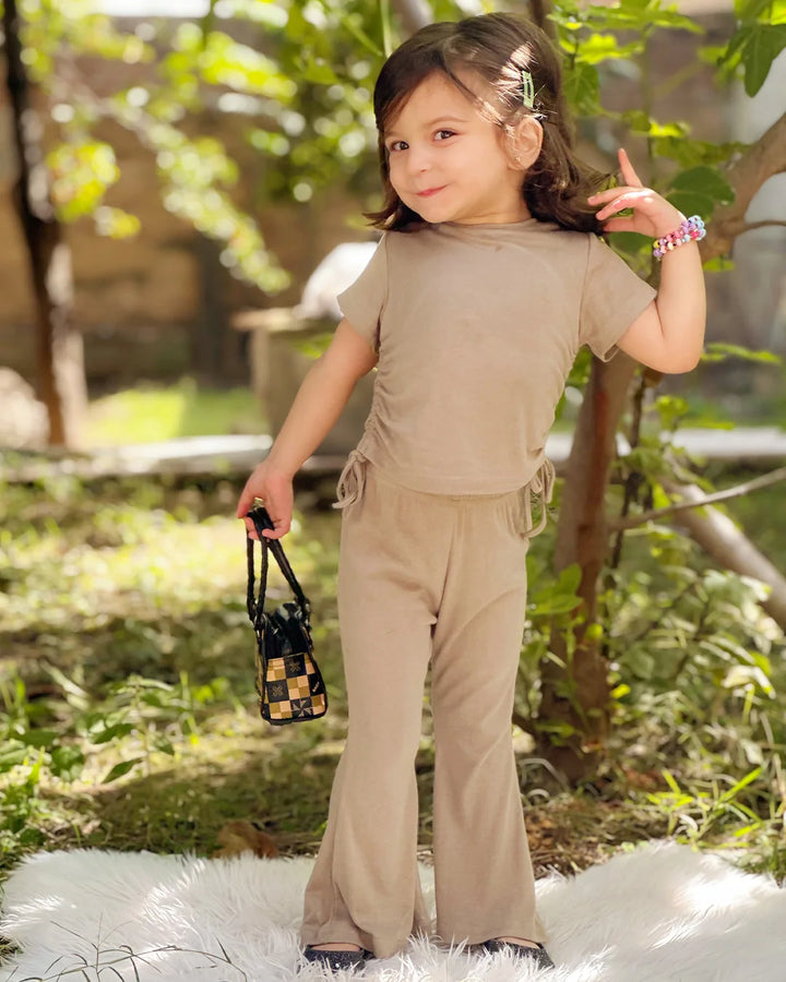 Young girl in a beige outfit standing outdoors with greenery in the background