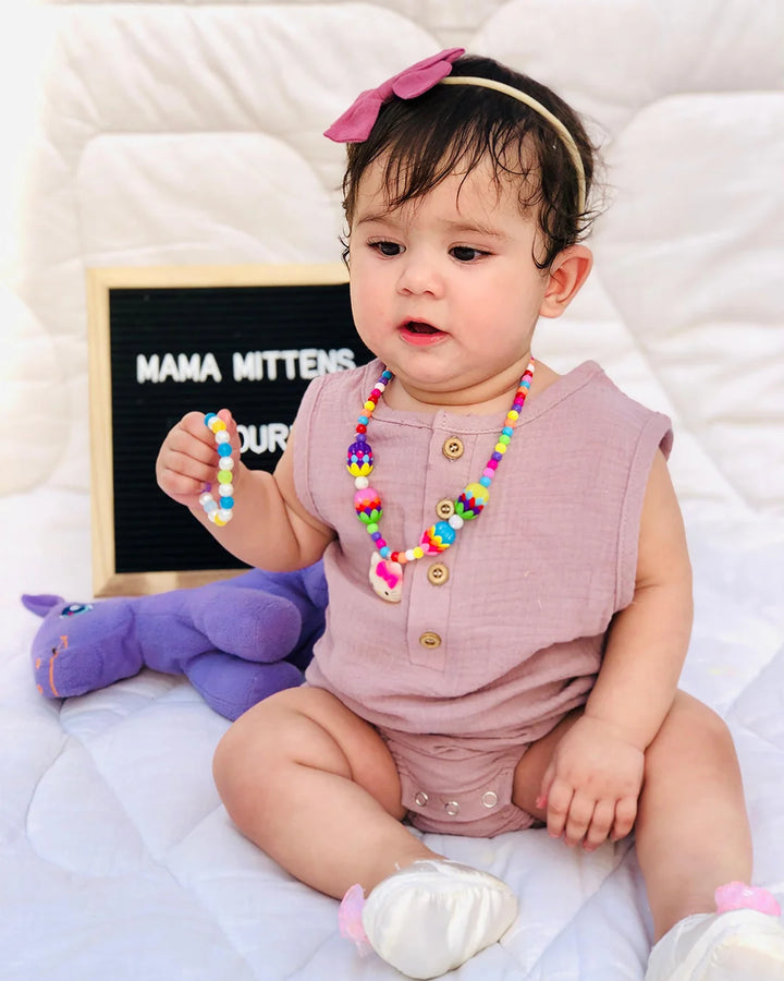 Baby wearing a pink outfit with colorful beads and a headband, sitting on a white surface.