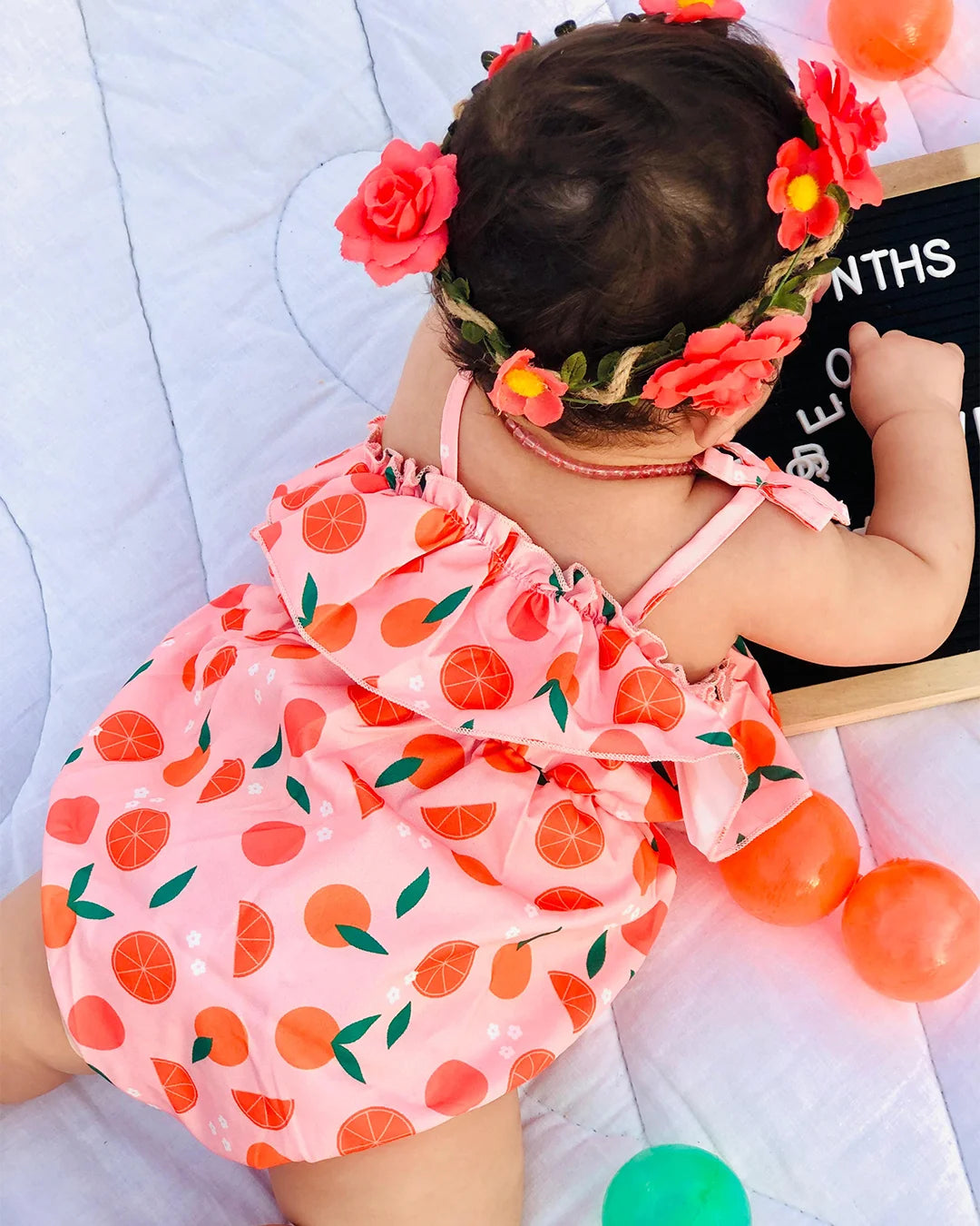 Baby in a pink outfit with orange fruit pattern, wearing a floral headband, on a light blue blanket.