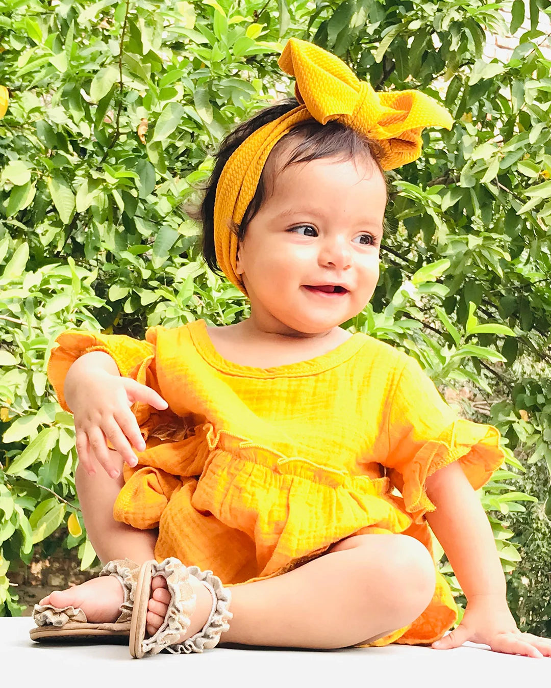 Baby in a yellow outfit with a large bow sitting outdoors among green leaves.