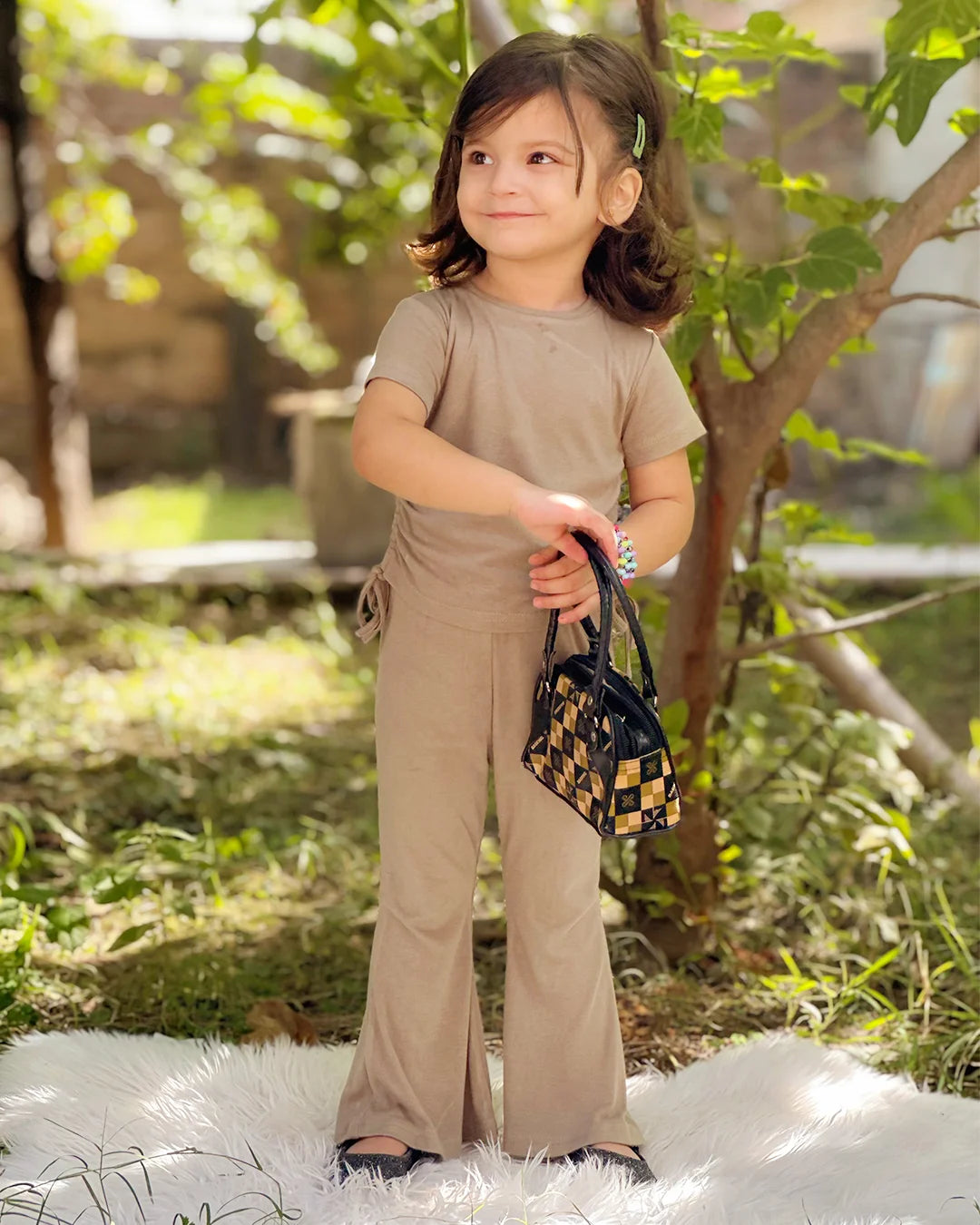 Child in beige outfit holding a small handbag outdoors