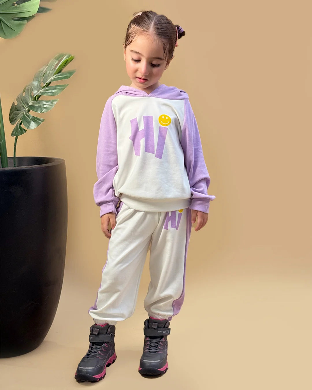 Child wearing a purple and white outfit with 'HI' text, standing next to a potted plant on a beige background
