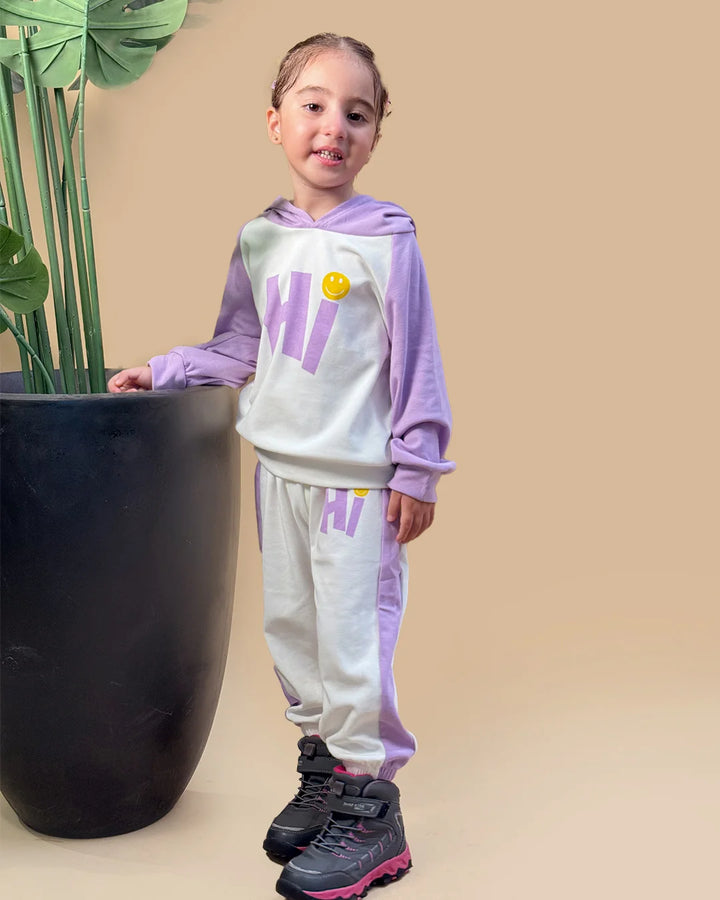 Child wearing a matching hoodie and sweatpants set with 'HI' print, standing next to a potted plant on a beige background.