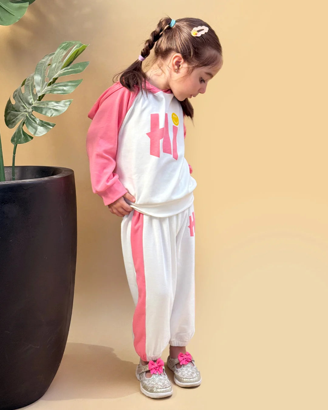 Child wearing a pink and white outfit with 'Hi' printed on it, standing next to a plant against a beige background.