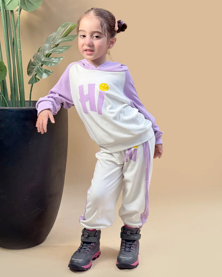 Child wearing a purple and white outfit with 'HI' printed on it, standing next to a potted plant.