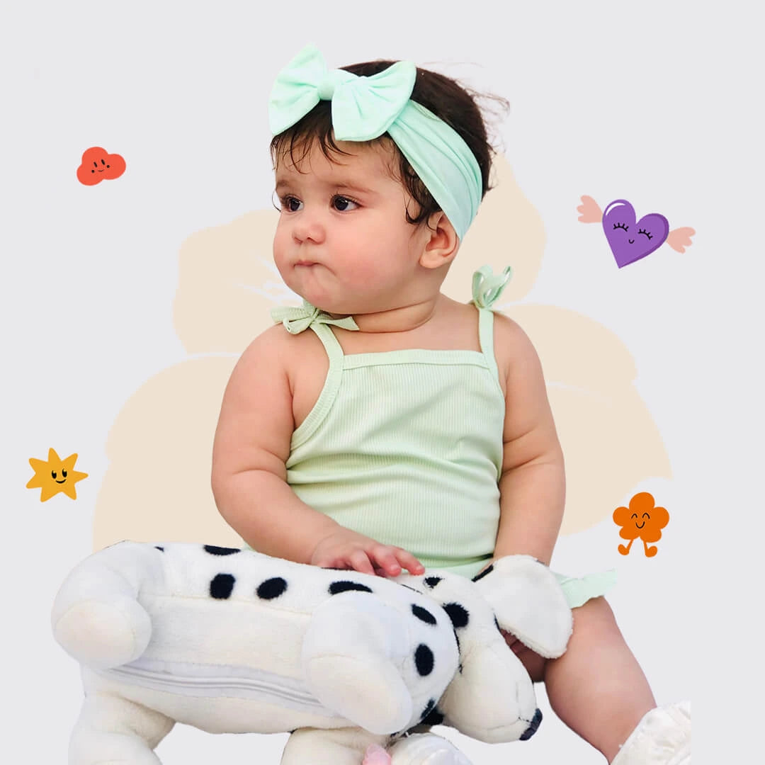 Baby in a light green outfit with a headband, sitting on a white cushion with a plush toy, surrounded by colorful stickers.