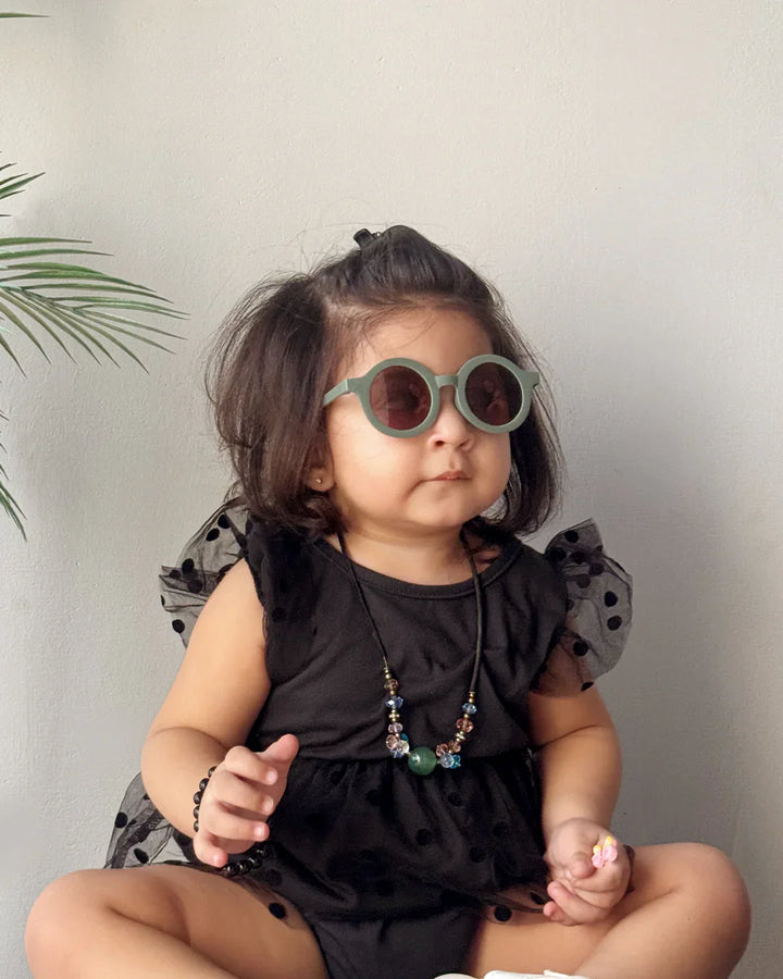 Child wearing a black outfit with ruffled sleeves and sunglasses, sitting against a neutral background.