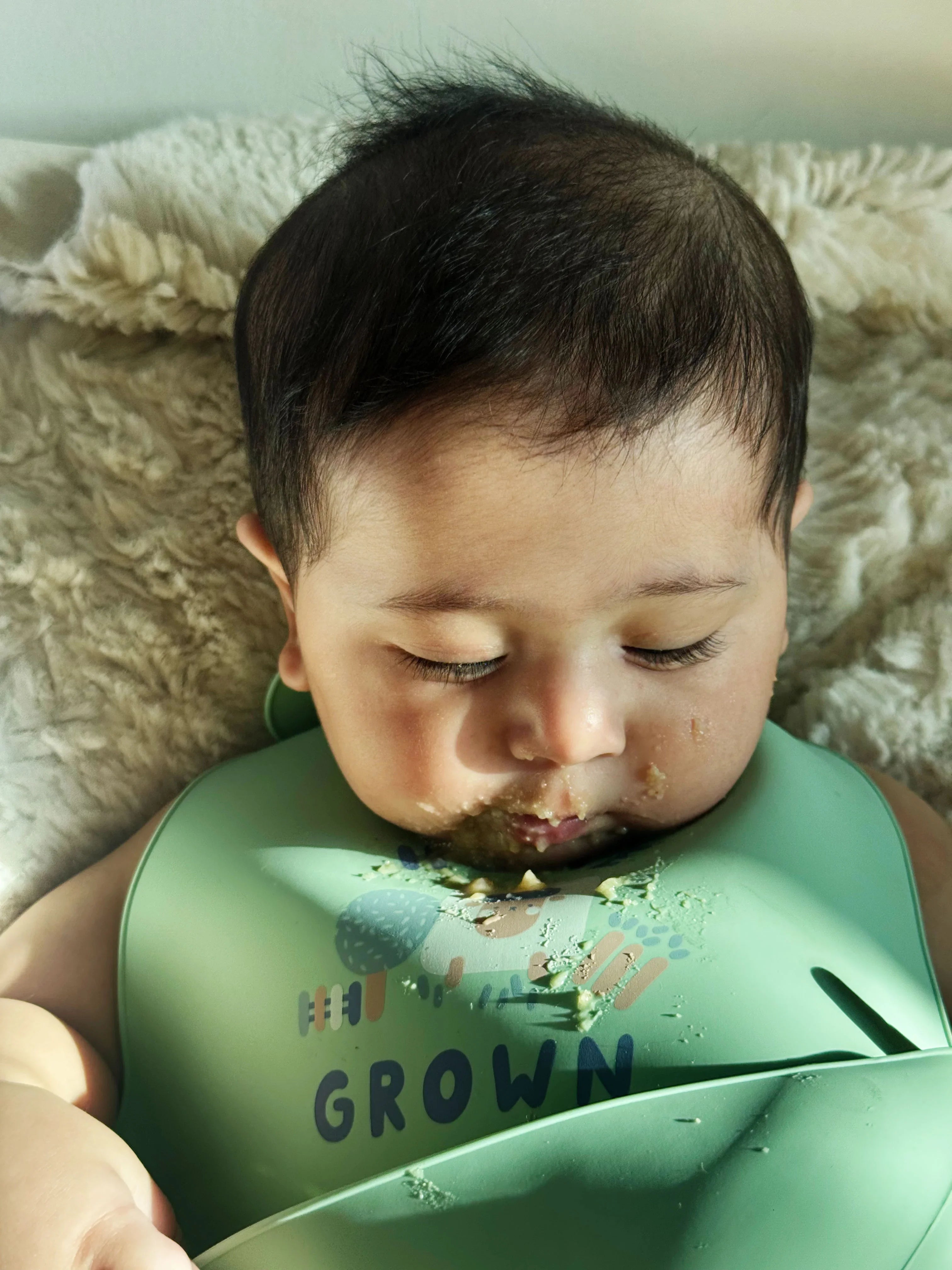 Baby wearing a green bib with 'Happy to be Grown' text, sitting on a soft surface.