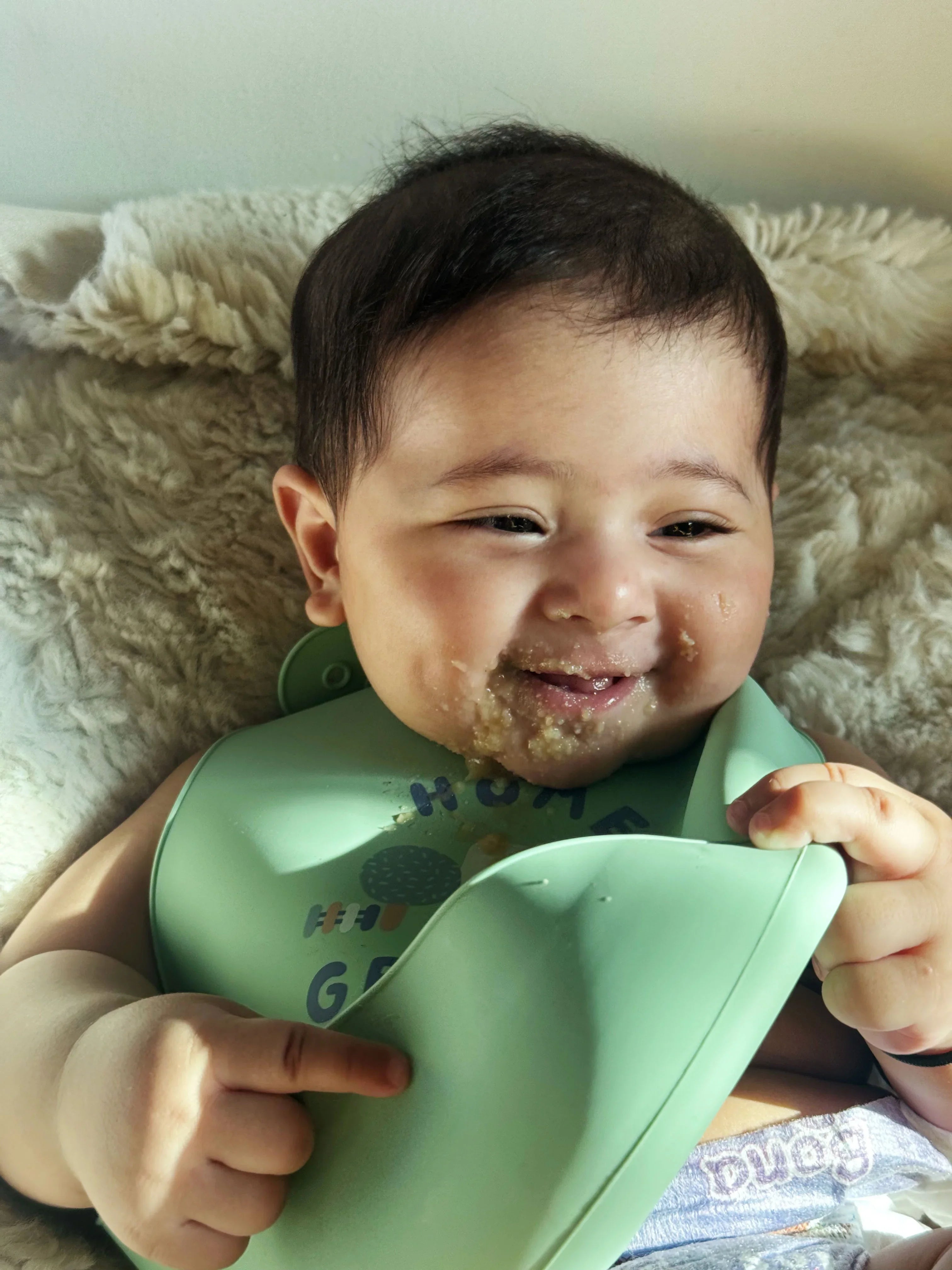 Baby wearing a green bib with food on it, lying on a soft surface.
