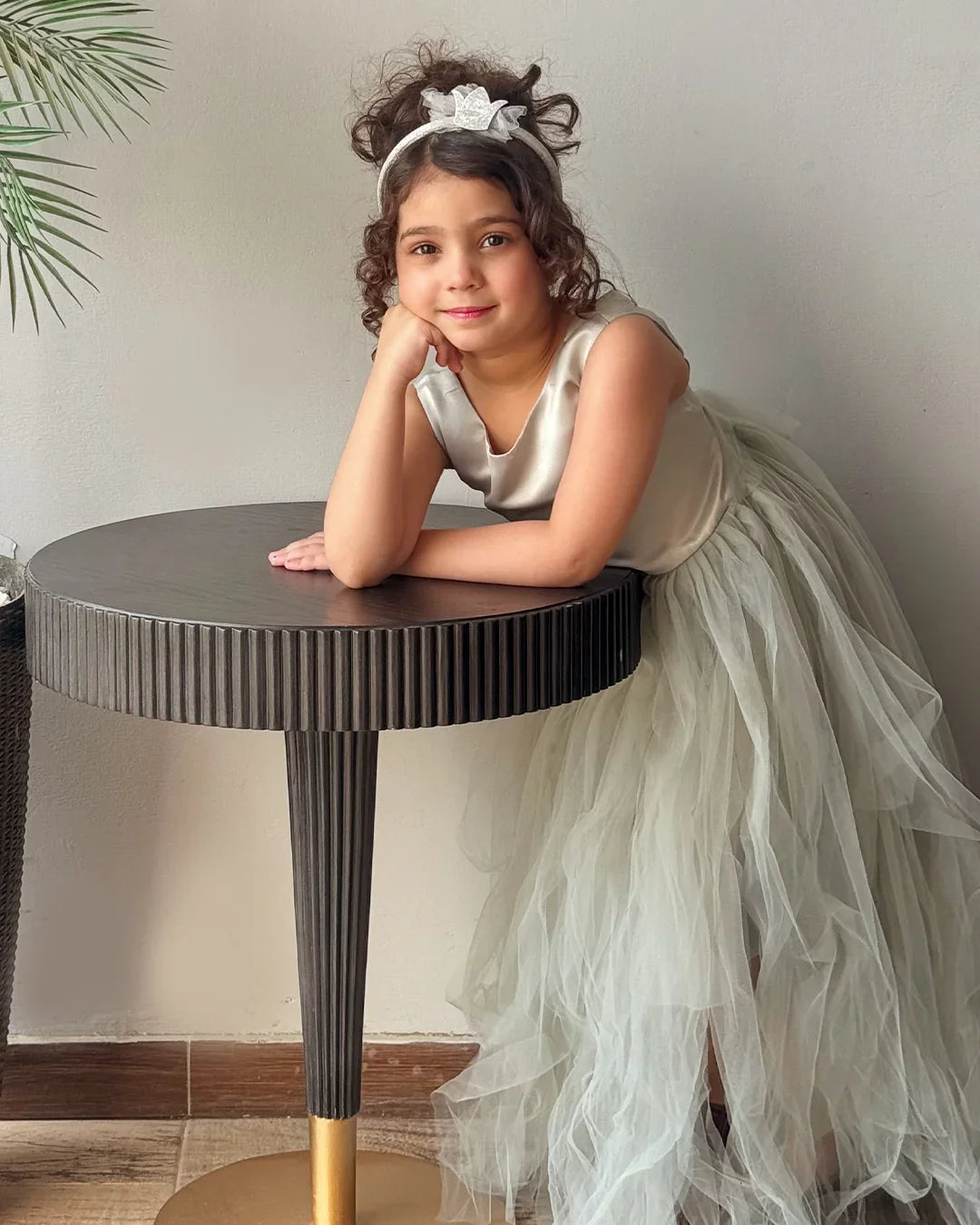 Young girl in a white dress with a tiara leaning on a round table indoors.