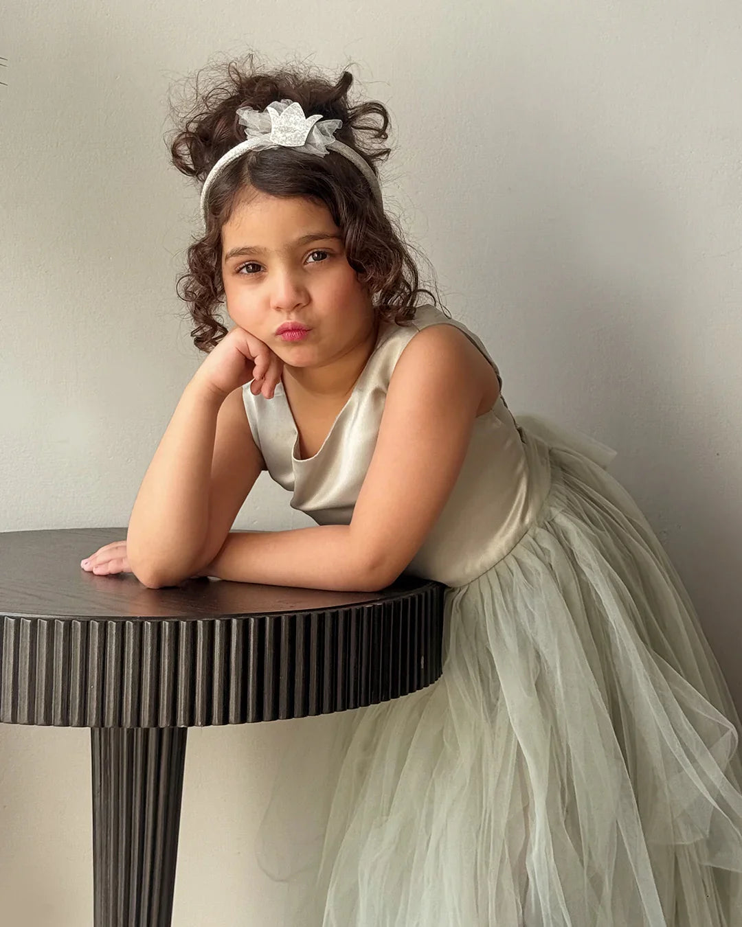 Young girl in a white dress with a headband sitting on a table against a plain background