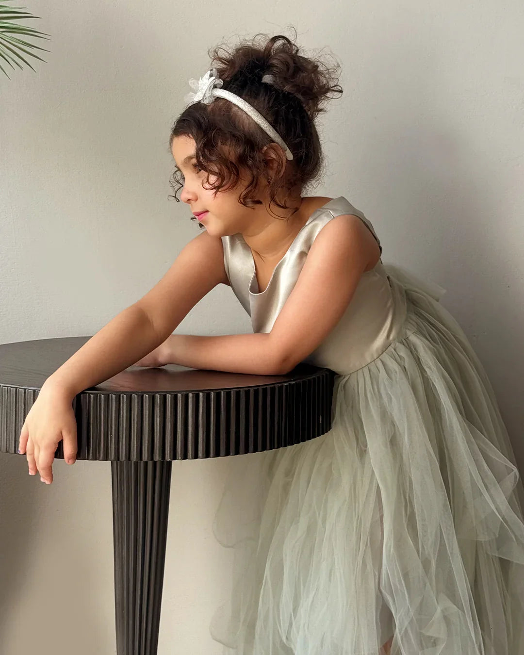 Woman in a white dress with a headband leaning on a table against a plain background
