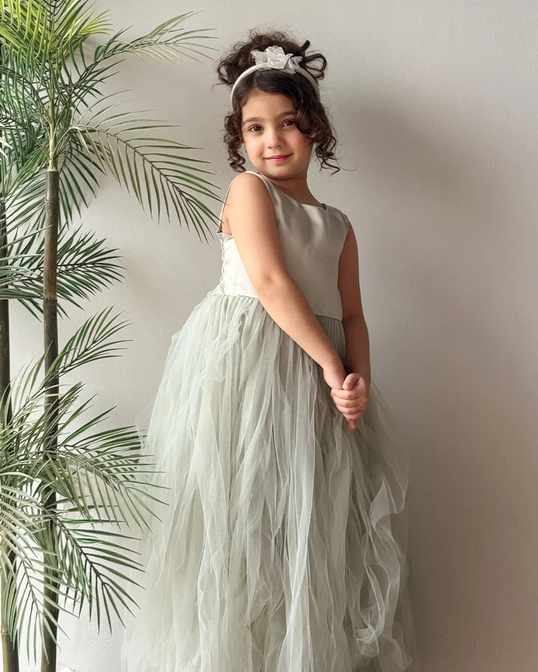 Young girl in a light green dress standing next to a plant against a plain wall.