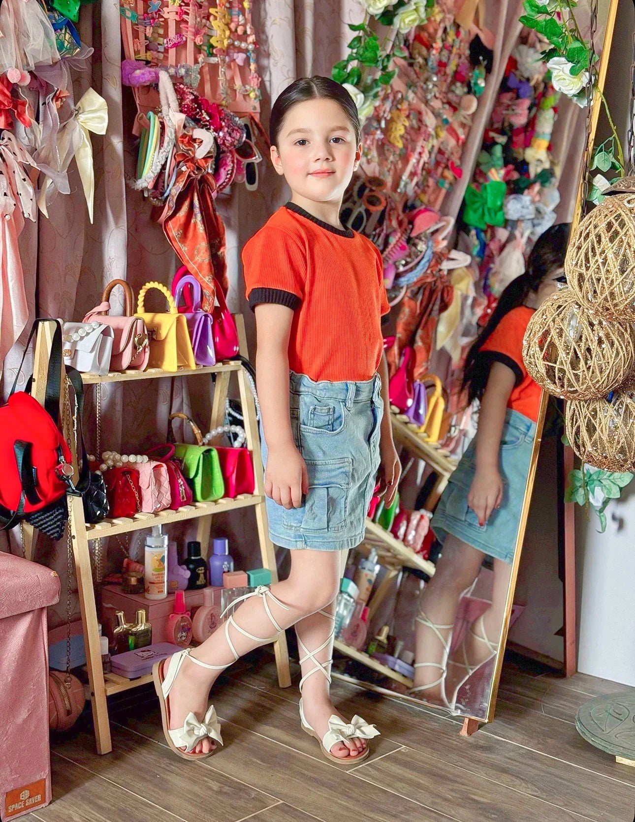 Young girl in an orange shirt and denim shorts standing in a colorful store.