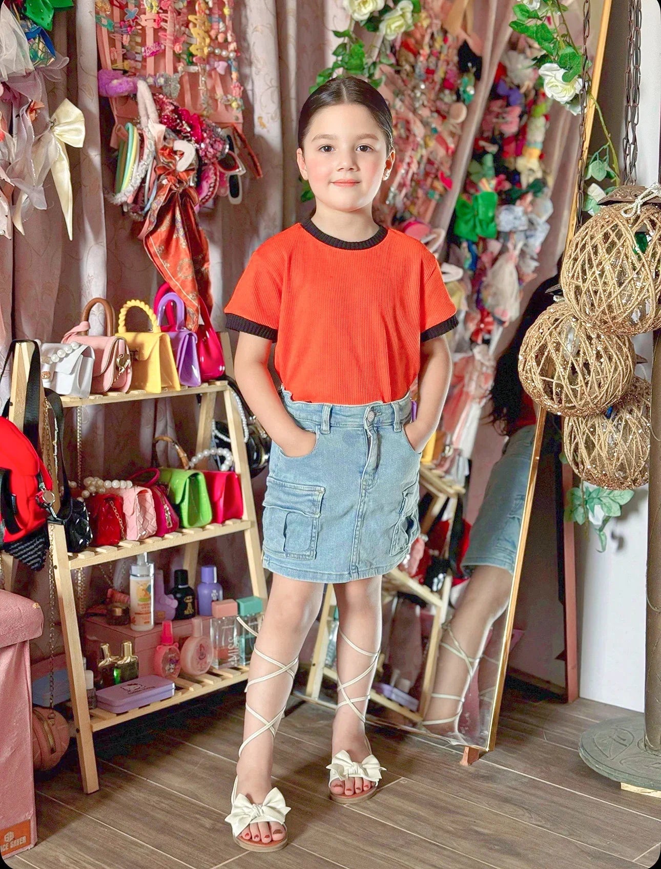 Young girl in a colorful store setting wearing an orange shirt and denim skirt.