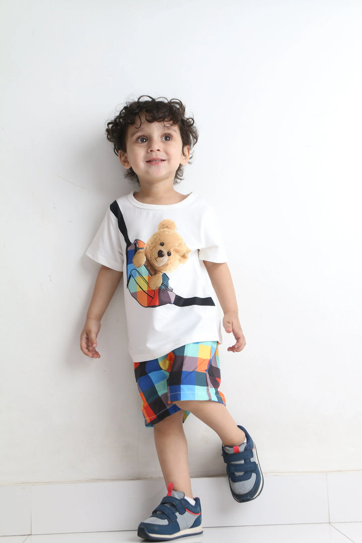 Child wearing a white t-shirt with a teddy bear design and colorful shorts on a white floor.