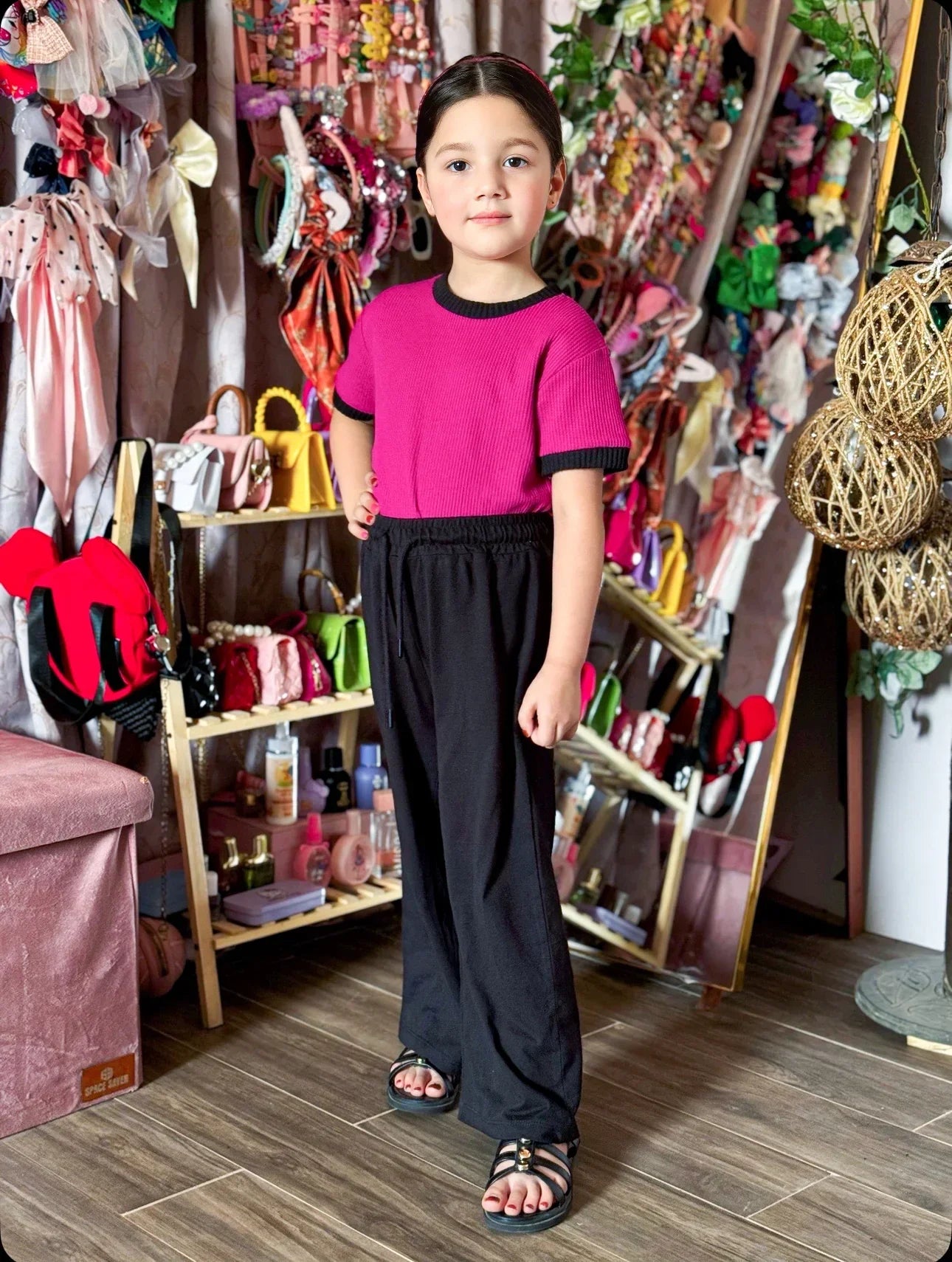 Child wearing a pink top and black pants standing in a store with various items on shelves.